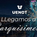 Una toma aérea nocturna que muestra un paisaje urbano con edificios de apartamentos de ladrillo a la izquierda y montañas al fondo. En el centro-derecha, una gran torre de reloj holográfica está hecha de líneas de luz azul cyan entrelazadas, con un reloj digital y una pantalla de texto en la parte superior, rodeada por una espiral de partículas de luz doradas y fragmentos. Un parque y carreteras están en el primer plano.