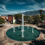 Vista aérea panorámica y de estilo cinematográfico de la Plaza las Heroínas en Mérida, Venezuela. En el centro destaca una gran fuente circular con chorros de agua activos, rodeada de caminerías, árboles verdes y edificios locales, con la imponente cordillera de los Andes de fondo bajo un cielo azul con nubes dispersas.